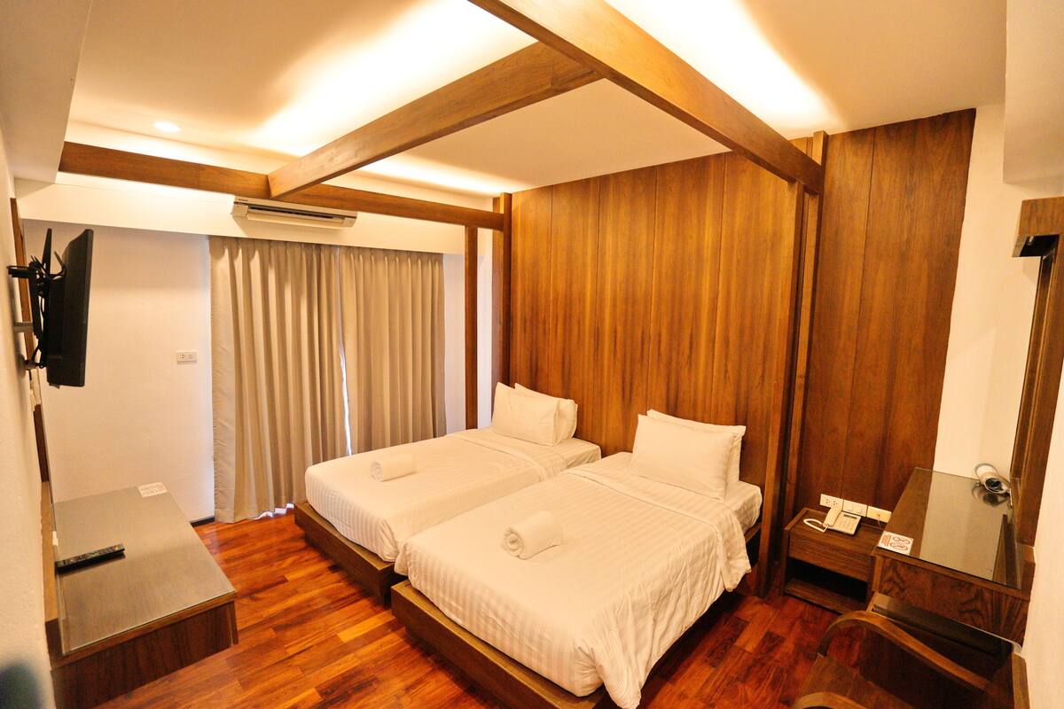 A hotel room with two beds, wooden flooring, a flat-screen TV, a bedside table, and a mirror.