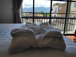 Un letto rifatto con coperta e cuscini bianchi, una finestra vista edifici e montagne, e una tenda a sinistra.