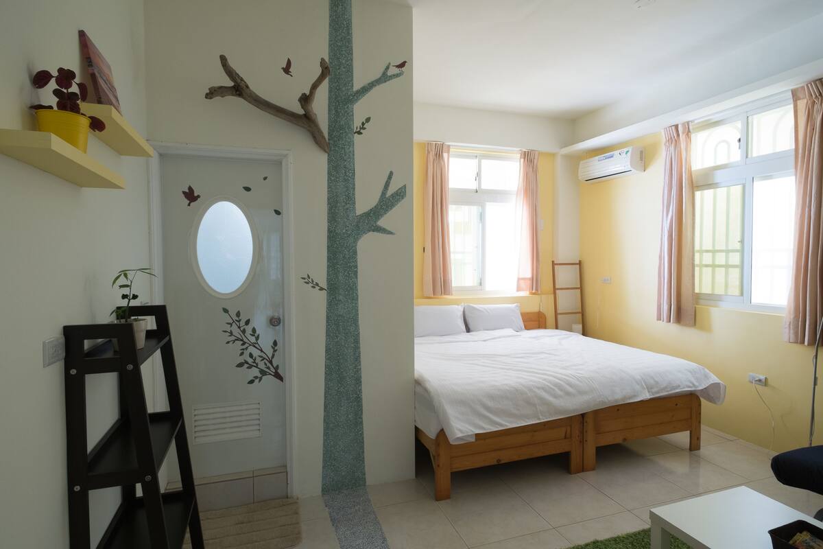 A bedroom with a wooden bed, a small desk, a chair, a ladder, and a window with curtains.