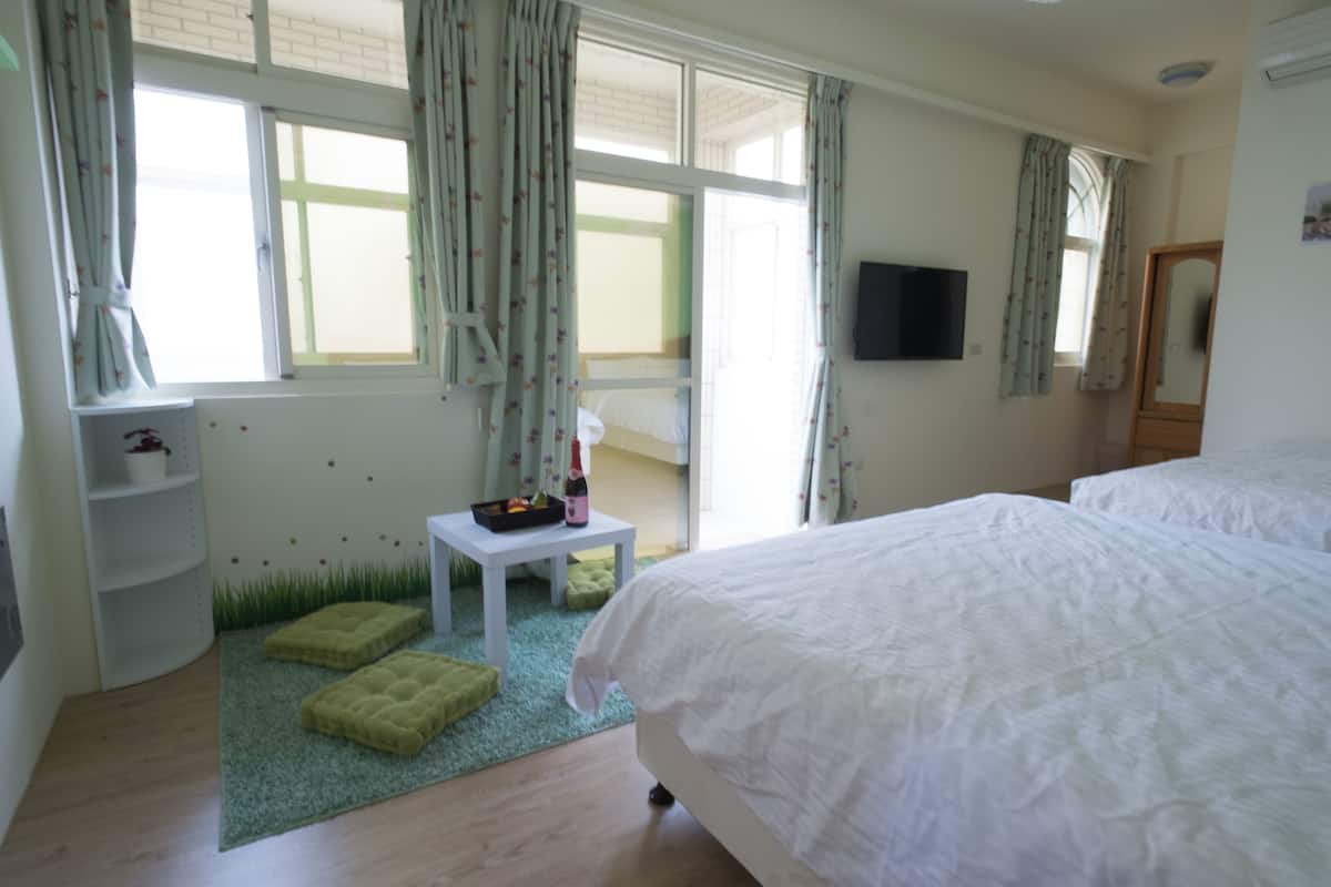 A bedroom with a large bed, a small table with a bottle and a bowl, a green rug, and a window with curtains.