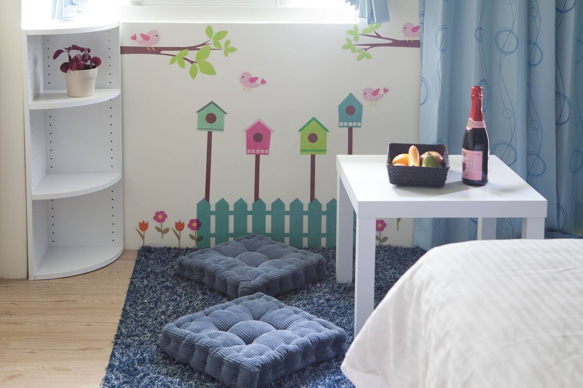 A child's room with a white bed, a small white table with a bottle and fruit basket, a blue rug, and a wall with bird and flower decals.