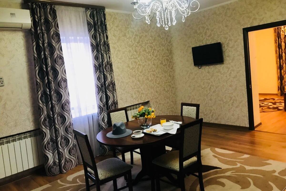 A dining area with a round table, four chairs, a chandelier, a flat-screen TV, and patterned curtains.