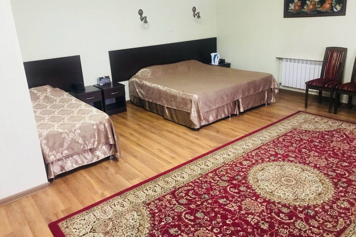 A hotel room with two beds, a wooden floor, a red and beige patterned rug, a nightstand with a lamp, and a chair.