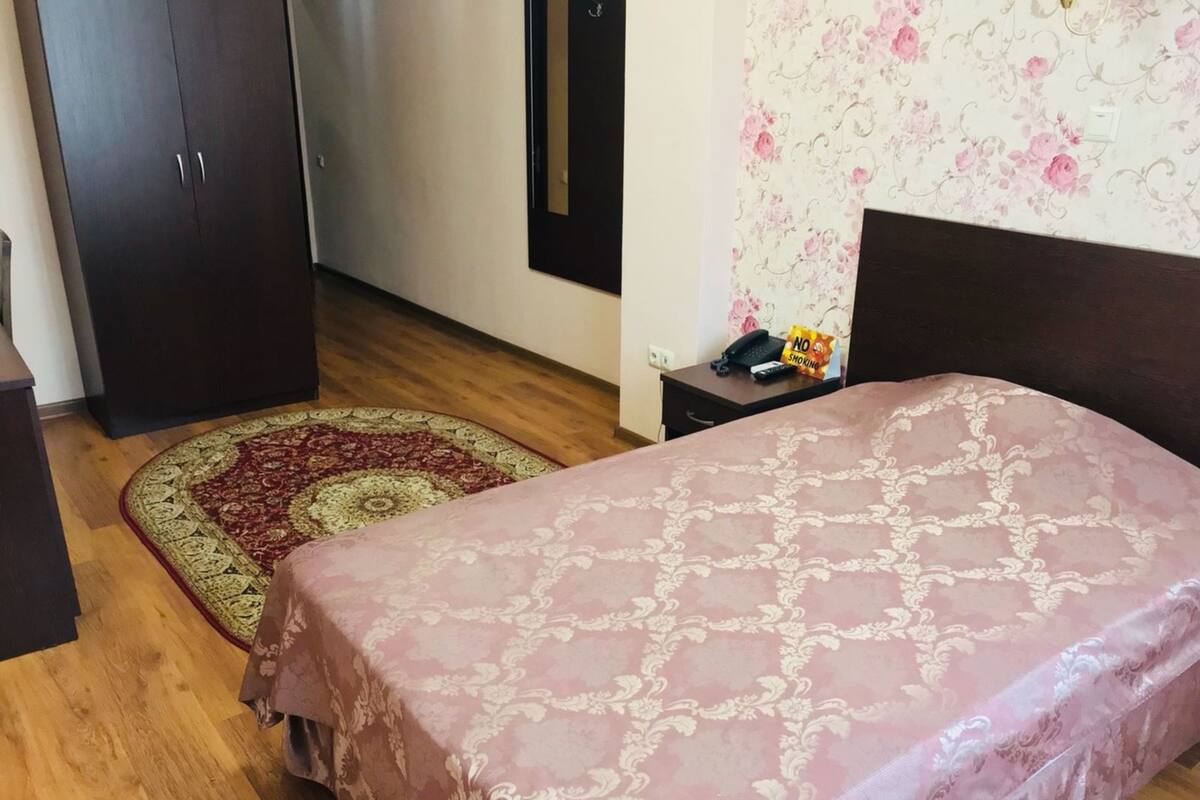 A bedroom with a bed, a nightstand, a desk, a wardrobe, and a patterned rug.