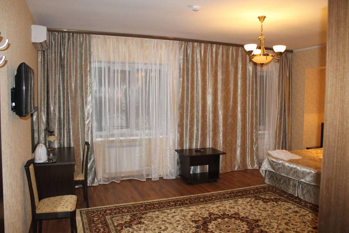 A hotel room with a wooden floor, a bed with a white bedspread, a small table with a lamp, a chair, a television mounted on the wall, and a chandelier.