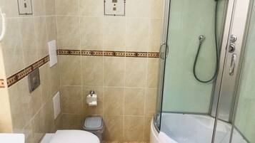 Pieni kylpyhuone, jossa on wc, roskakori, lasiovilla varustettu suihku ja pyyheteline.