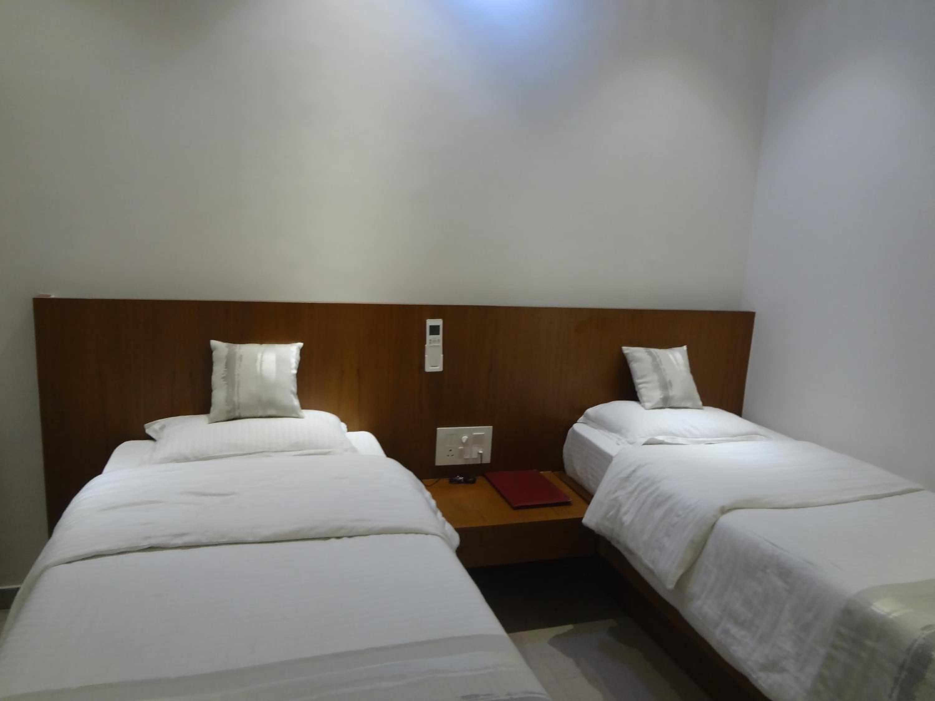 A hotel room with two single beds, a wooden headboard, a nightstand with a red book, and a wall-mounted thermostat.