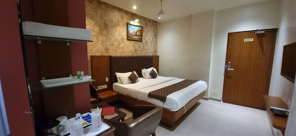 Habitación de hotel con cama, escritorio, silla, repisa con artículos de aseo, un cuadro en la pared y una puerta de salida.
