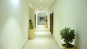 Un pasillo luminoso y bien iluminado, con piso y paredes blancos e impecables, que conduce a una puerta al final. Hay plantas en macetas a lo largo de los laterales del pasillo.