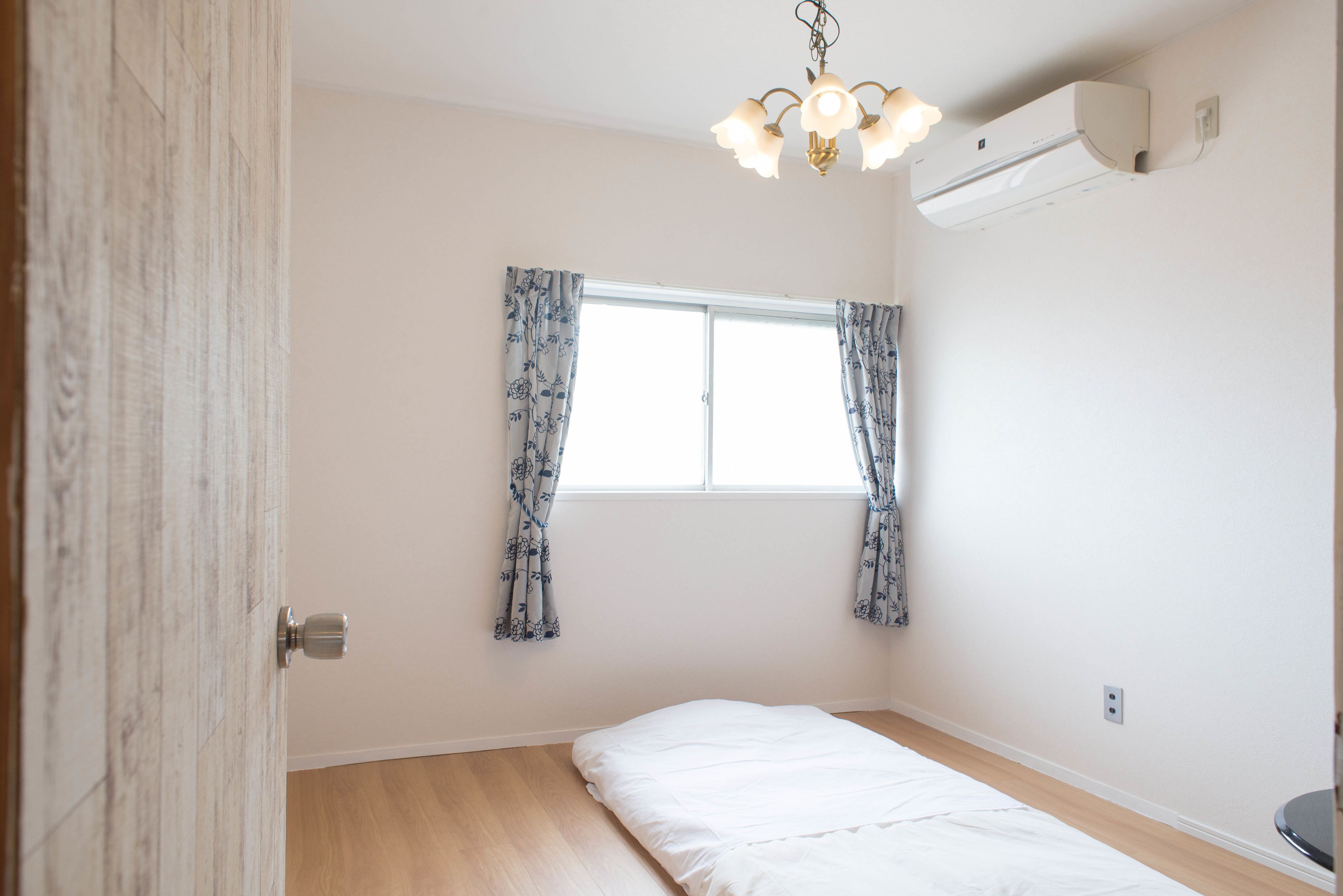 Een kamer met een bed, een raam met gordijnen, een plafondlamp en een airconditioning.