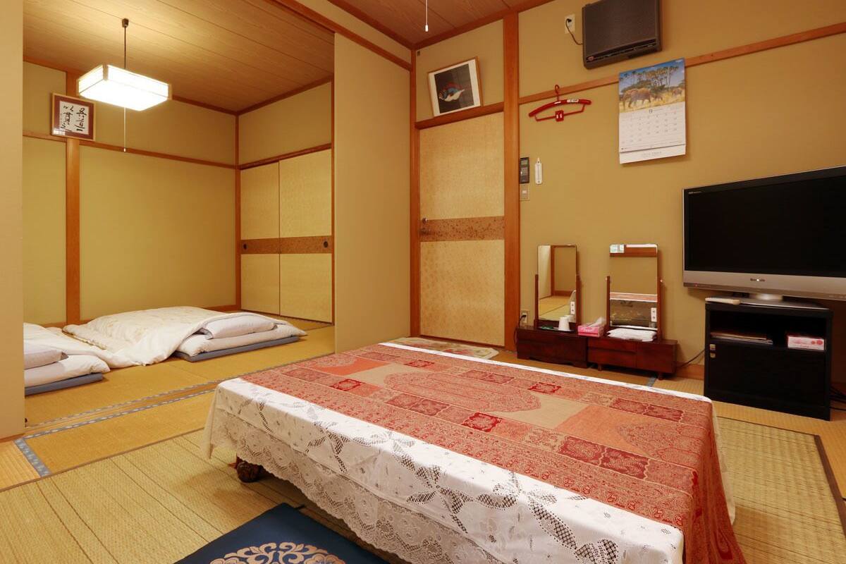 A traditional Japanese-style room with tatami flooring, a bed with a red and white patterned bedspread, a television, and a calendar on the wall.