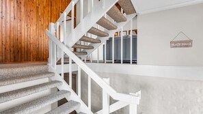A staircase with white railings and carpeted steps leading to a room with a wooden wall and a sign reading 'BEACH HOUSE'.