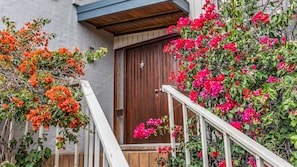 Una porta de fusta amb una barana blanca al davant, envoltada de plantes de buganvília de colors vives.