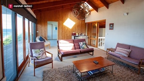 A cozy living room with a wooden ceiling, a large window, and a view of the outdoors. - Ocean Pearl - Bunga St 61 (BERMAGUI)