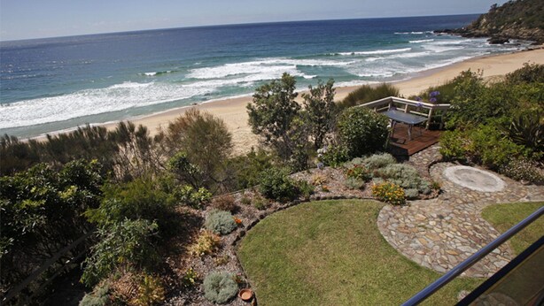 A coastal landscape with a beach, ocean, and a patio area with a table and chairs. - Ocean Pearl - Bunga St 61 (BERMAGUI)