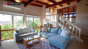 A spacious living room with a large window, a staircase, and a bookshelf. - Ocean Pearl - Bunga St 61 (BERMAGUI)