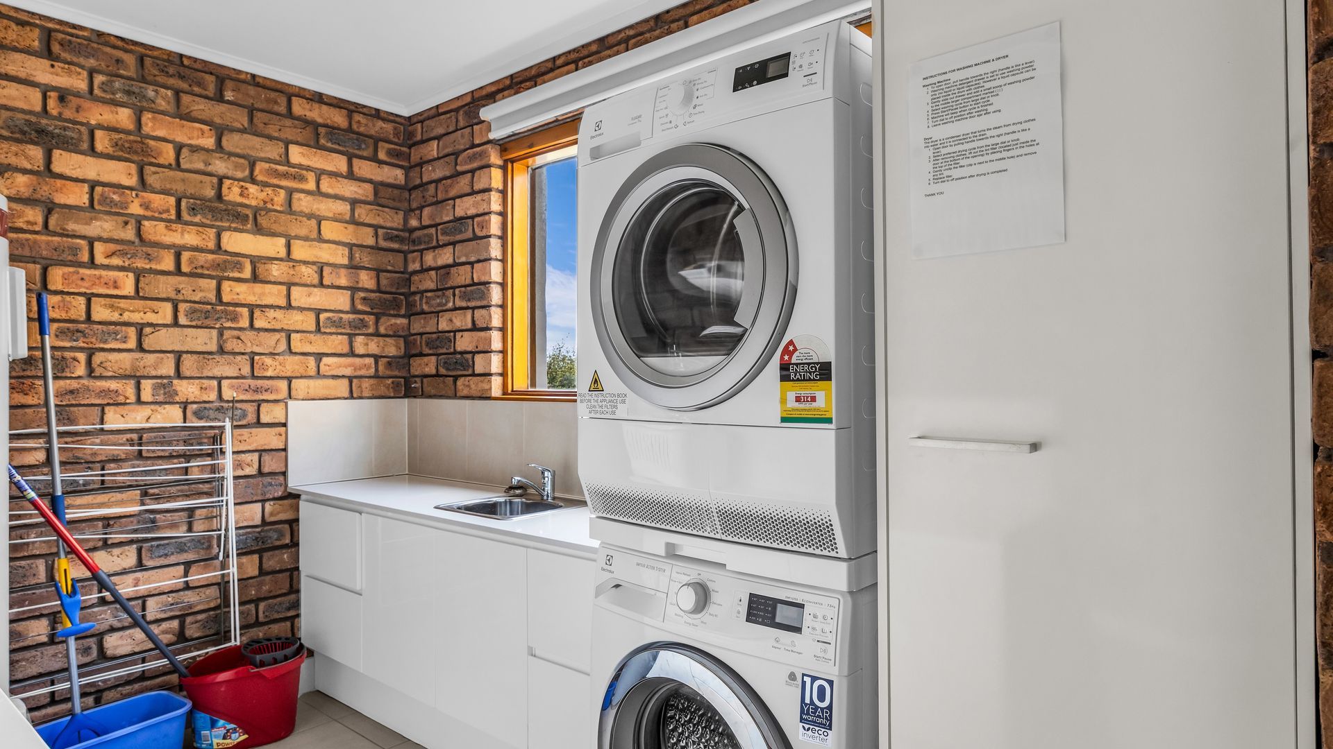 Een wasserette met een bakstenen muur, een gestapelde wasmachine en droger, een wasbak en een raam.