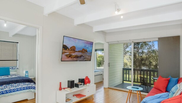 A modern bedroom with a large bed, a TV, a small table, and a balcony with a view of the outdoors. - Eight On The Lake - Fairhaven Point Way 8 (WALLAGA LAKE)