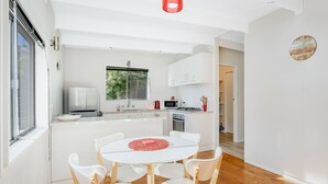 A modern kitchen with white cabinetry, a dining table with four chairs, and a red decorative item on the table. - Eight On The Lake - Fairhaven Point Way 8 (WALLAGA LAKE)