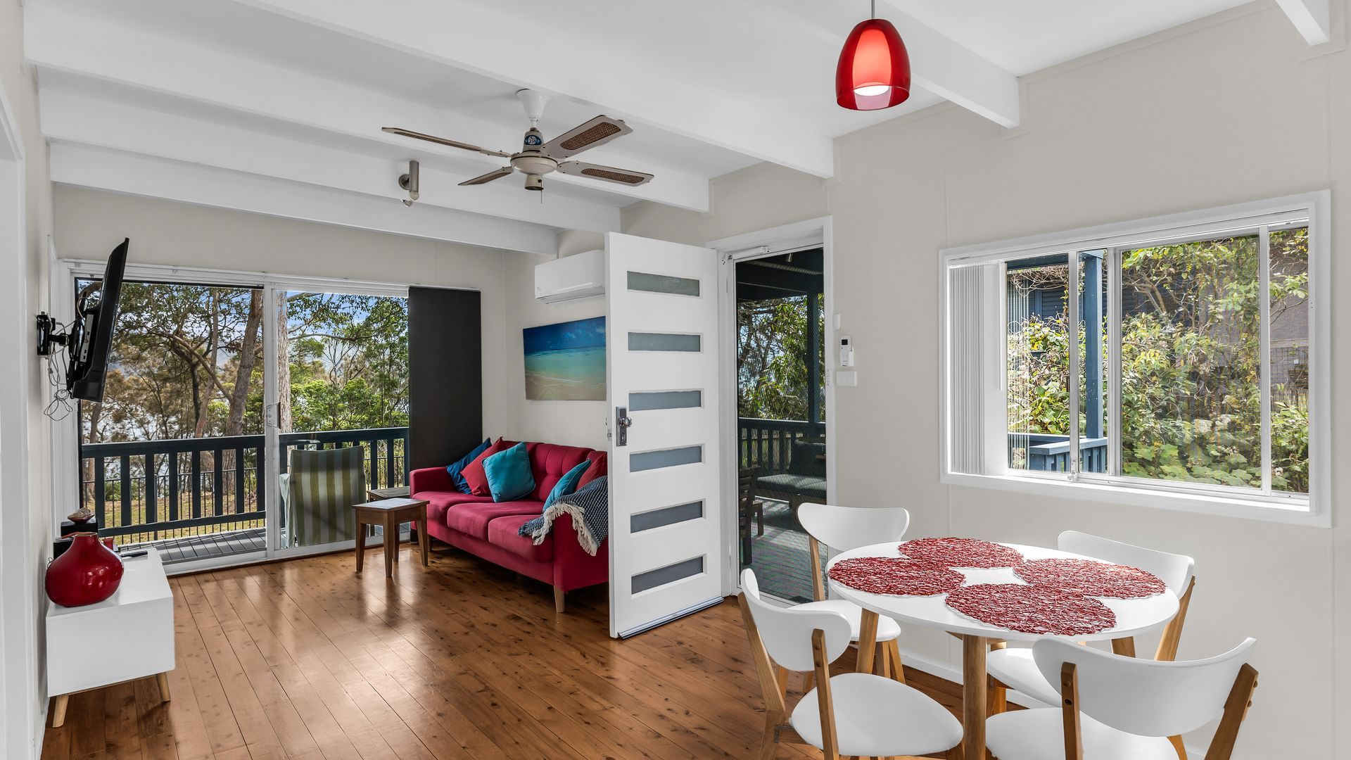 A spacious room with a red sofa, white chairs, a dining table with a red table runner, a ceiling fan, and large windows with a view of trees.