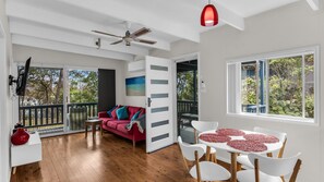 A spacious room with a red sofa, white chairs, a dining table with a red table runner, a ceiling fan, and large windows with a view of trees.