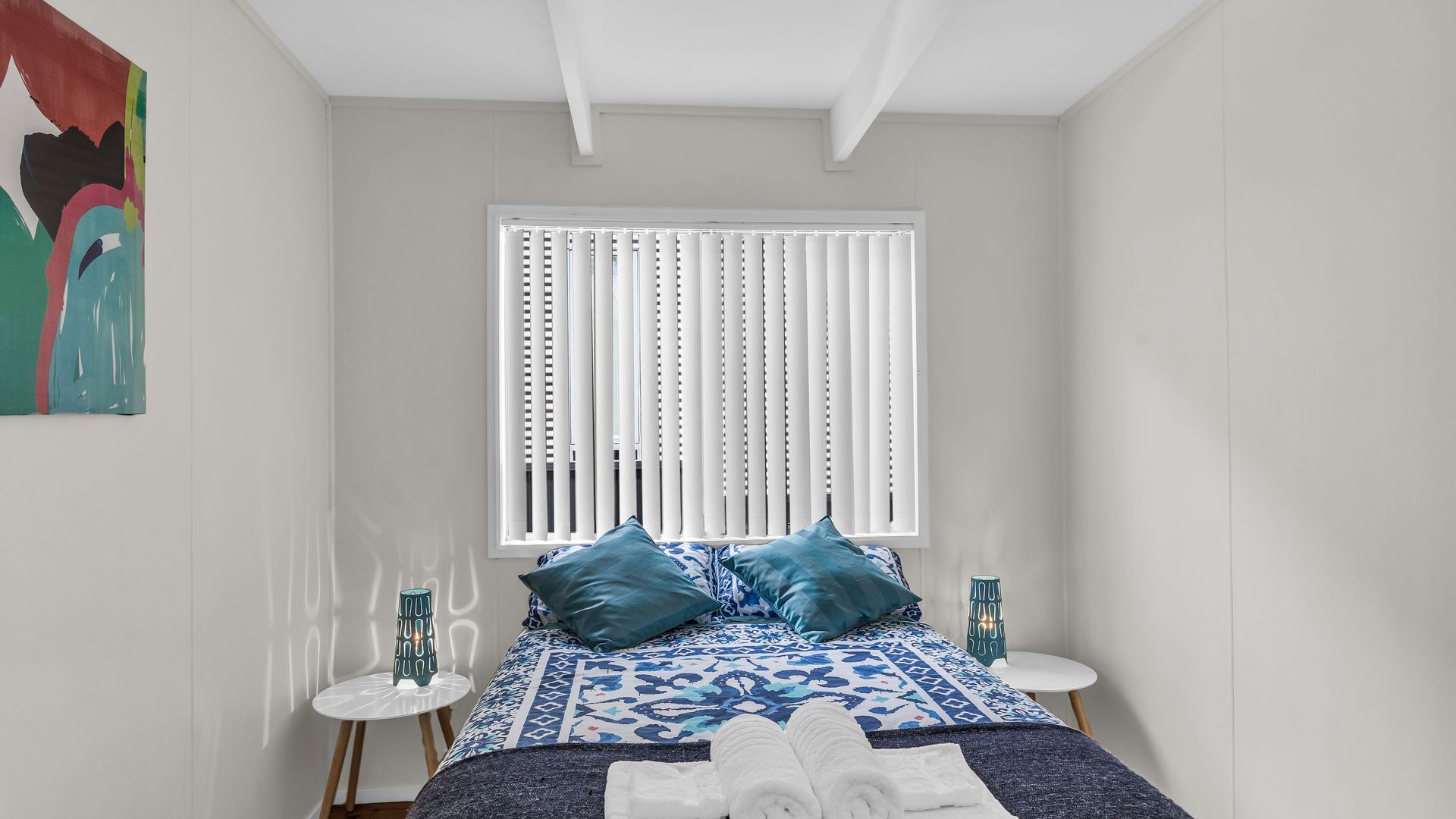 Une chambre à coucher avec un lit, deux tables de chevet, une fenêtre avec des stores et un tableau au mur.