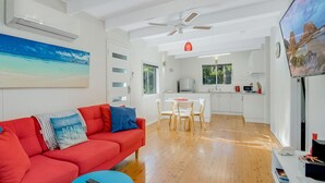 A modern living room with a red sofa, a painting of a beach, a dining area with a table and chairs, and a kitchen in the background. - Eight On The Lake - Fairhaven Point Way 8 (WALLAGA LAKE)