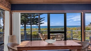Une terrasse en bois avec une table et des chaises, offrant une vue sur un paysage côtier avec des arbres et la mer.