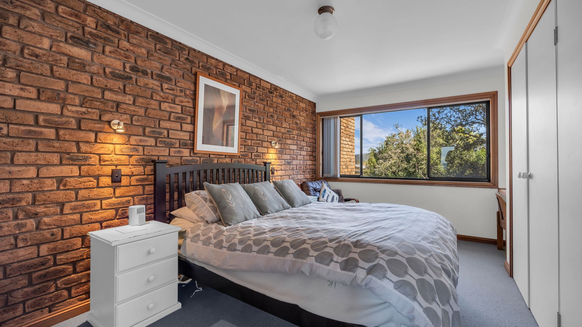 Ein Schlafzimmer mit Backsteinwand, einem Bett mit gemusterten Bettbezügen, einem Nachttisch, einem Fenster mit Ausblick und einem gerahmten Bild an der Wand.