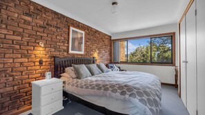 Une chambre avec un mur en briques, un lit avec une literie à motifs, une table de chevet, une fenêtre donnant sur un paysage agréable et un tableau encadré accroché au mur.