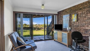 A room with a sliding glass door leading to a garden, a television on a wooden cabinet, a chair, and a desk with a chair. - Breakwater 1 - Narira Street 1 (Bermagui)