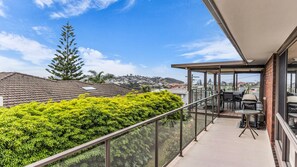 A balcony with a glass railing, a table with chairs, and a view of a cityscape. - Mumbulla House - Mumbulla St 22a (BERMAGUI)