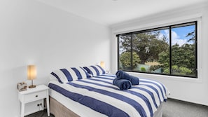 A bedroom with a large window, a bed with striped bedding, a bedside table with a lamp, and a view of trees outside. - Mumbulla House - Mumbulla St 22a (BERMAGUI)