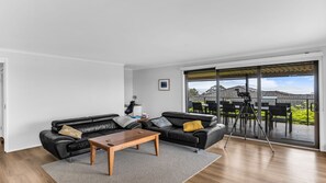 A modern living room with a black leather sofa, a wooden coffee table, and a large sliding glass door leading to an outdoor patio with a camera on a tripod. - Mumbulla House - Mumbulla St 22a (BERMAGUI)