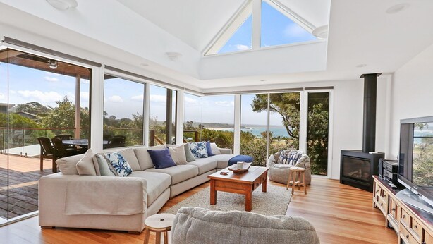 A spacious living room with a large sofa, a coffee table, and a television. The room has large windows offering a view of the sea and greenery. - Cuttagee Views - Cuttagee Beach Road 22 (BARRAGGA BAY)