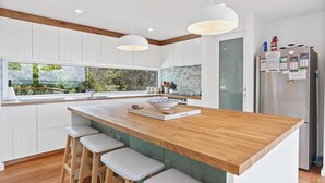 A modern kitchen with a wooden island, white cabinetry, and a large window offering a view of greenery. - Cuttagee Views - Cuttagee Beach Road 22 (BARRAGGA BAY)