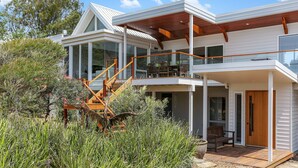 A modern two-story house with a wooden staircase leading to a balcony, surrounded by dense vegetation. - Cuttagee Views - Cuttagee Beach Road 22 (BARRAGGA BAY)