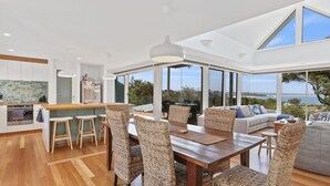 A modern kitchen with a central island, a dining area with wicker chairs, and a large sliding glass door leading to a balcony with a view of the sea. - Cuttagee Views - Cuttagee Beach Road 22 (BARRAGGA BAY)