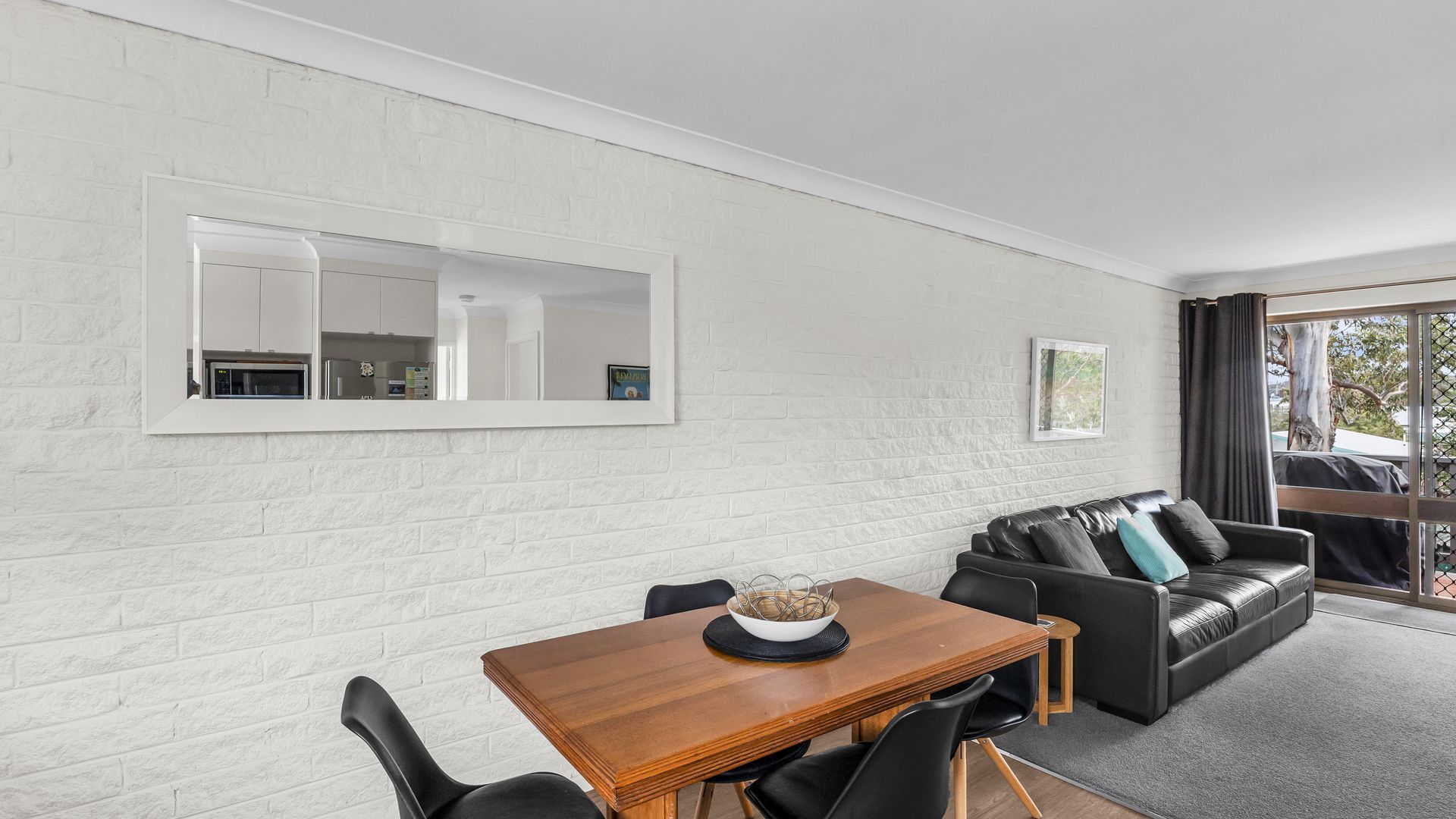 Un salon moderne comprenant un coin repas, des murs en briques blanches, un grand miroir et un canapé en cuir noir.