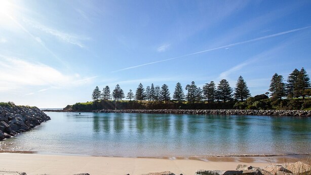 A beach with calm water, a rocky shoreline, and a line of trees in the background.