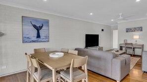 A modern living room with a sofa, a dining table with chairs, a flat-screen TV, and a ceiling fan.