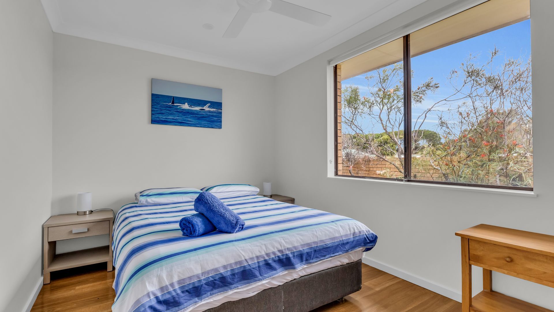Un dormitorio con cama, mesita de noche, ventana con vista a árboles y ventilador de techo.