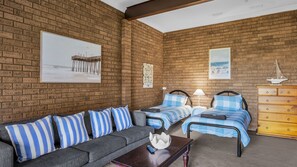 A room with a brick wall, a sofa, two beds with blue bedding, a wooden dresser, and artwork. - Beares Beach House - Mill Street 7 (BERMAGUI)