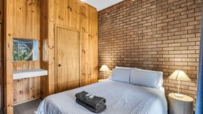 A room with a wooden panel wall, a brick wall, a bed with a checkered bedspread, two bedside lamps, and a shelf with a book. - Beares Beach House - Mill Street 7 (BERMAGUI)