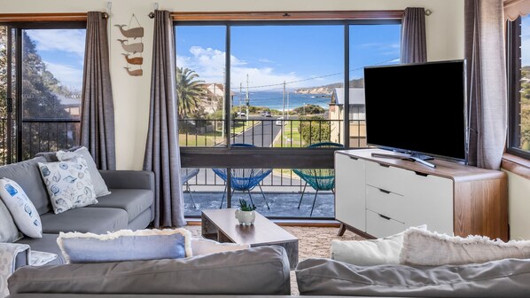 A living room with a sofa, a coffee table, and a TV. There is a balcony with chairs and a view of the sea.