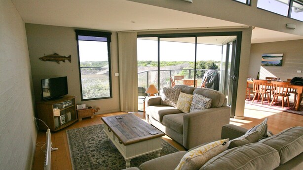 A spacious living room with a large window, a television, a wooden coffee table, and a sofa. - Moorhen Cove 3 - Unit 3 Wallaga Lake Rd 15 (BERMAGUI)