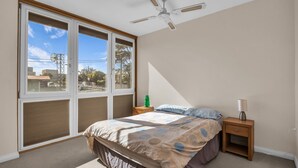Une chambre avec une grande fenêtre, un lit, une table de chevet, une lampe et un ventilateur de plafond.
