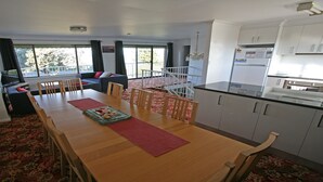 A modern kitchen with white cabinets, a dining area with a wooden table and chairs, and a living area with a sofa and a TV. - Gull Cottage - Pacific Drive 7 (BERMAGUI)