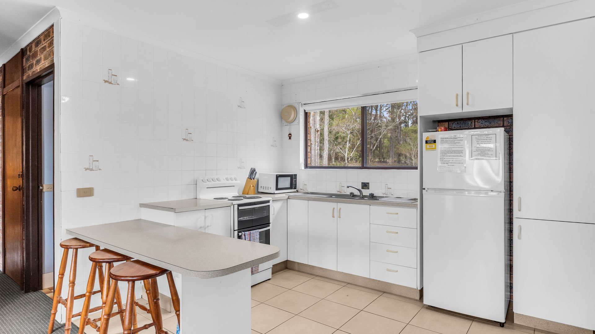 A modern kitchen with white cabinets, a built-in oven, a microwave, and a refrigerator. There is a window providing a view of trees outside.
