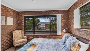 A bedroom with a large window, a bed, a chair, and a view of a building outside. - Cuttagee Beach Cottage 3 - Tathra Bermagui Rd 3521 (BARRAGGA BAY)